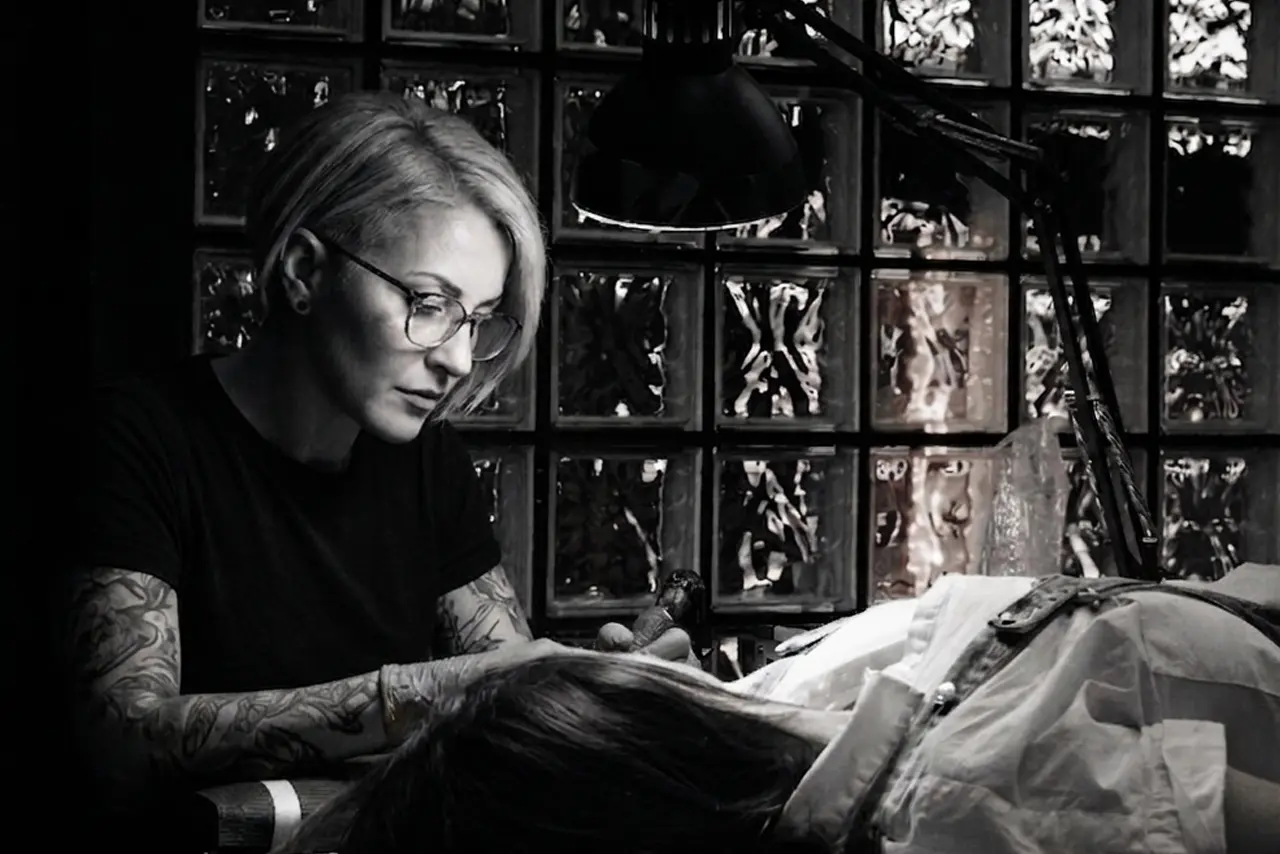 A professional female tattoo artist with glasses and tattooed arms focused on inking a client in a modern studio with a glass block wall background.