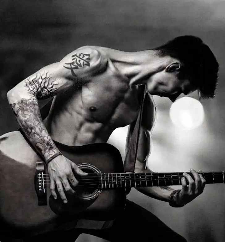 Shirtless man playing guitar featuring a bold black ink Chinese tattoo for Love on his right shoulder.